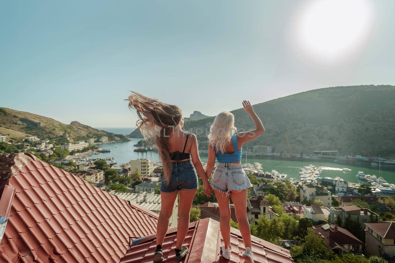 women standing on rooftop, enjoys town view and sea mountains. Peaceful rooftop relaxation. Below her, there is a town with several boats visible in the water. Rooftop vantage point