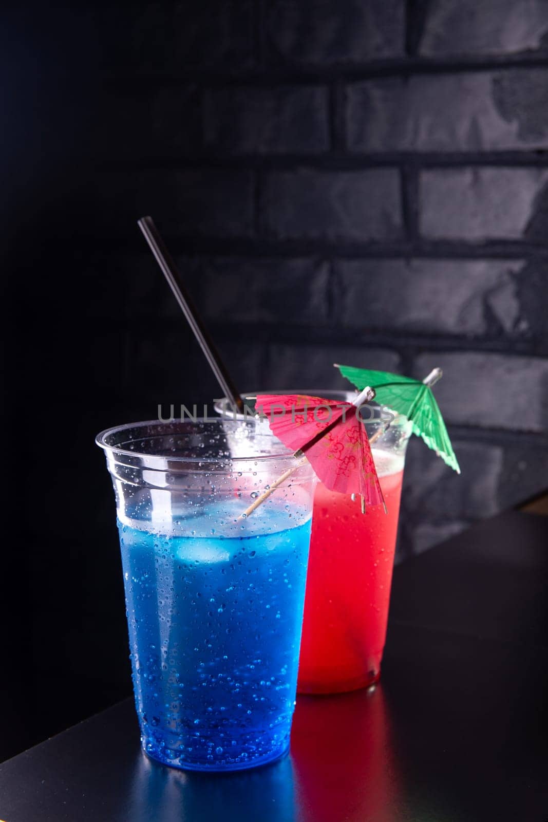 Red and blue lemonade with ice cubes in a plastic glasses decorated with an umbrella and straws on black wooden counter against a brick wall