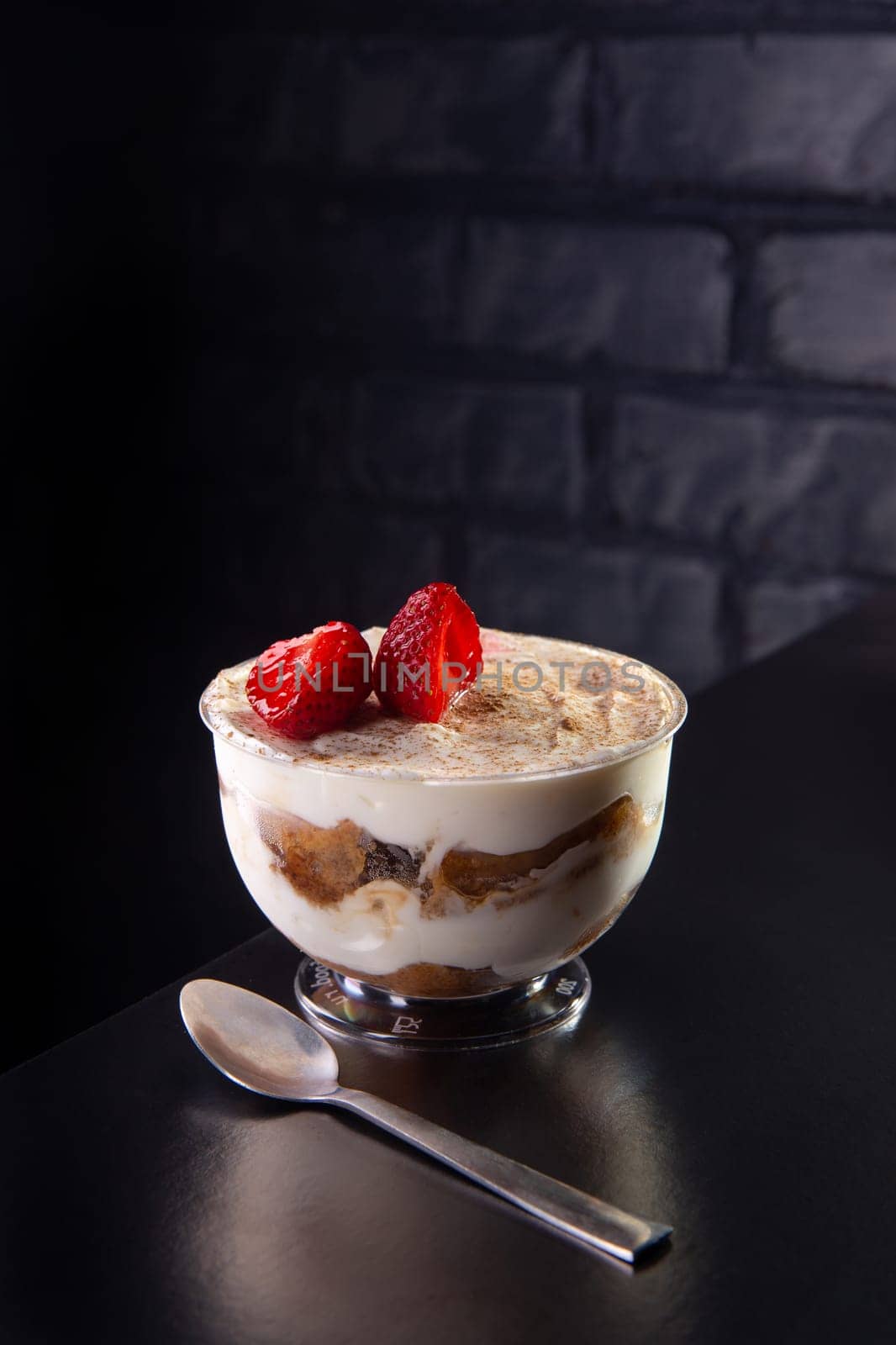 Tiramisu dessert in a glass bowl, decorated with coffee and strawberries on black table with brick wall behind