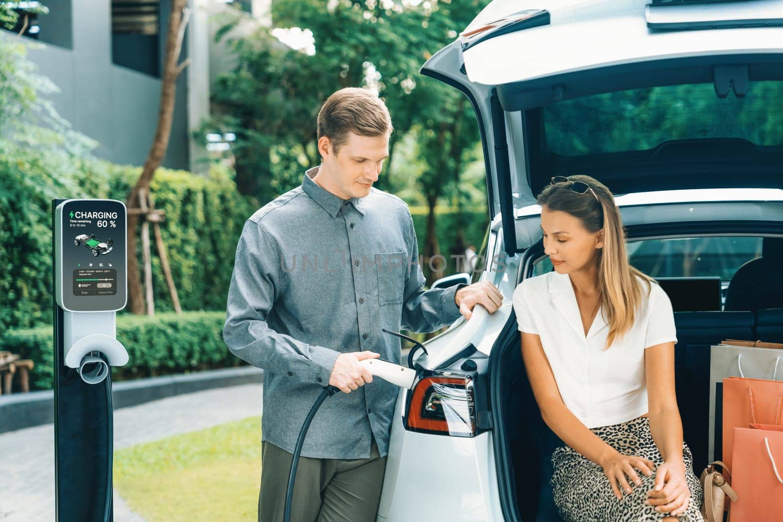 Young couple travel with EV electric car charging in green sustainable city outdoor garden in summer shows urban sustainability lifestyle by green clean rechargeable energy of electric vehicle innards