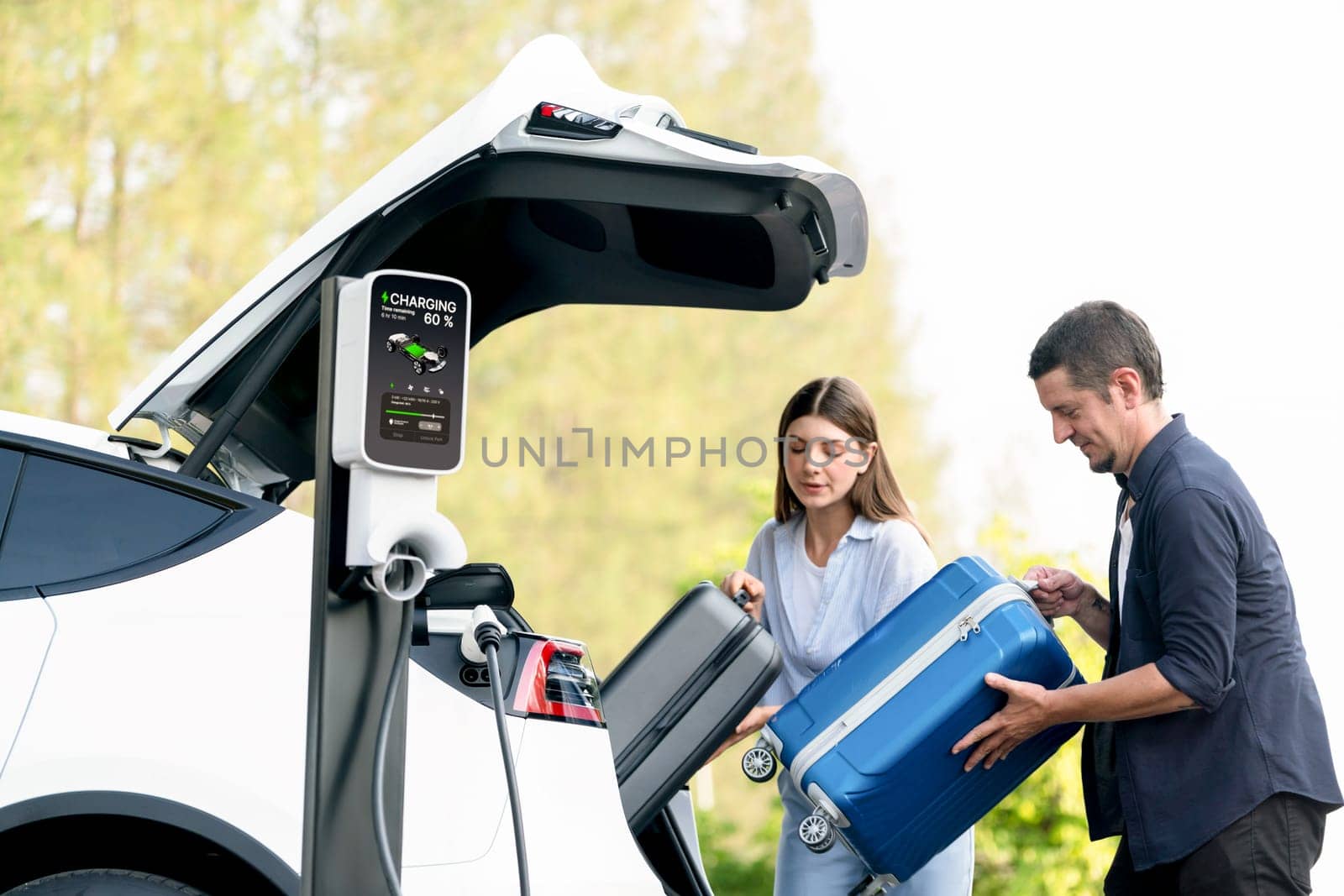Lovely young couple recharging battery for electric car while relaxing and sitting on the trunk during road trip travel EV car in autumnal forest. Eco friendly travel on vacation during autumn. Exalt