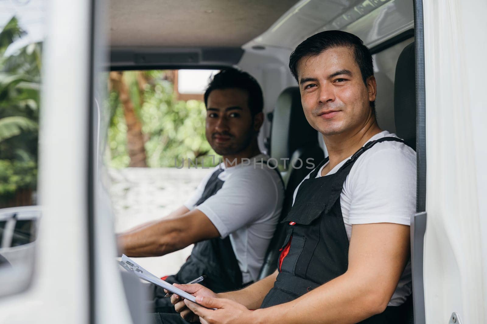 Ethnicity-diverse mover team driving a truck for relocation service. Two smiling workers in uniform delivering furniture with confidence. Transportation and teamwork emphasized. Moving Day Concept.