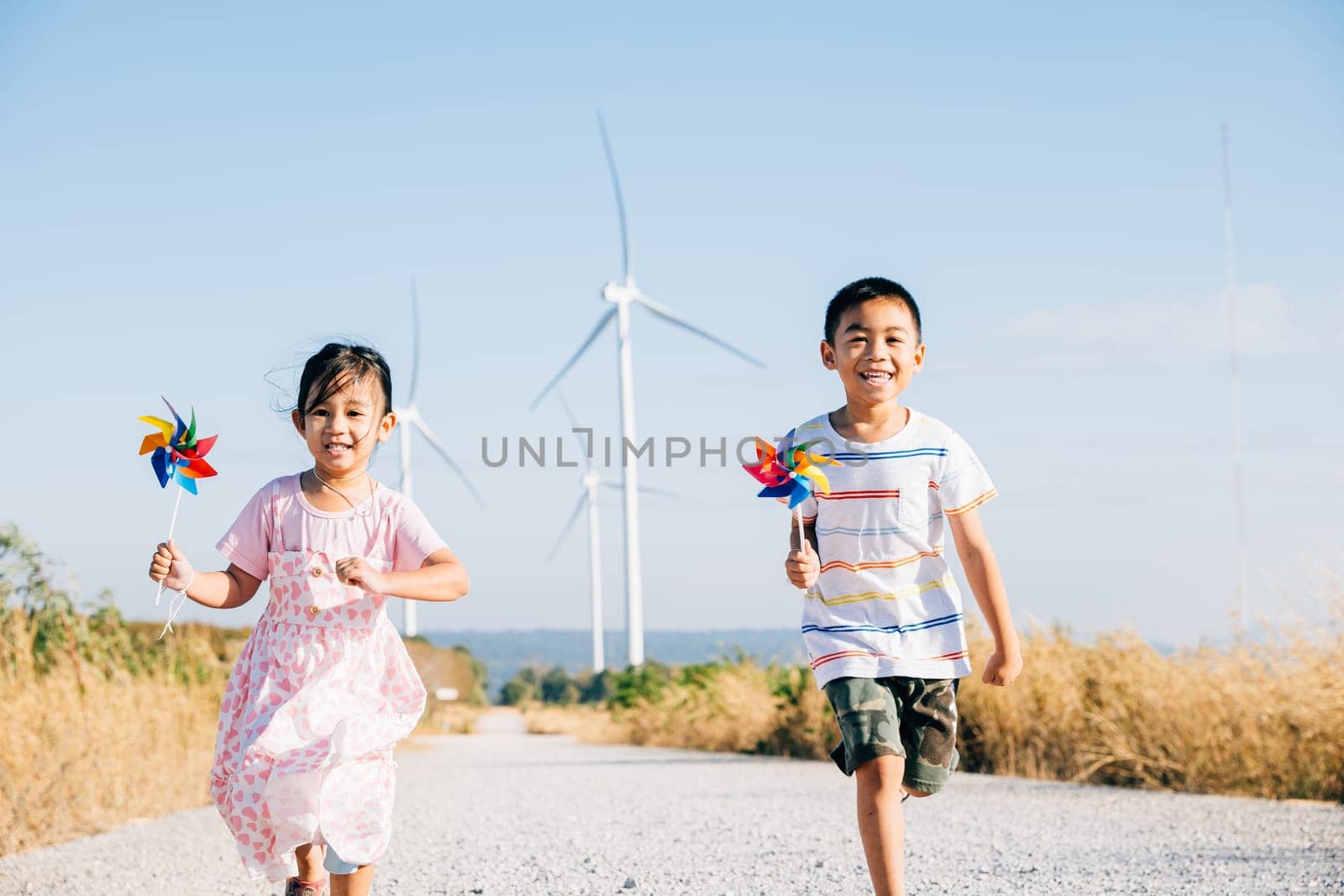 Brother and sister run by windmills holding pinwheels. Kids' joy near turbines represents a future of clean energy. Smiling children carefree and happy engage with sustainable technology.