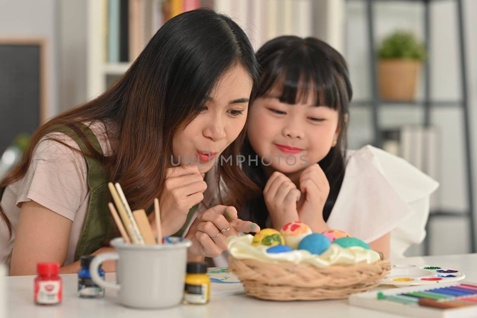 Little child girl and mother preparing for Easter holiday at home together, painting eggs in living room.