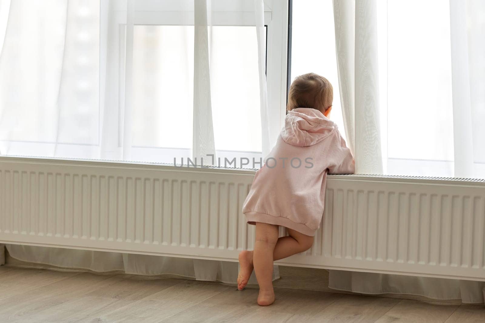 Little child girl standing at window and looking outside. Back view.