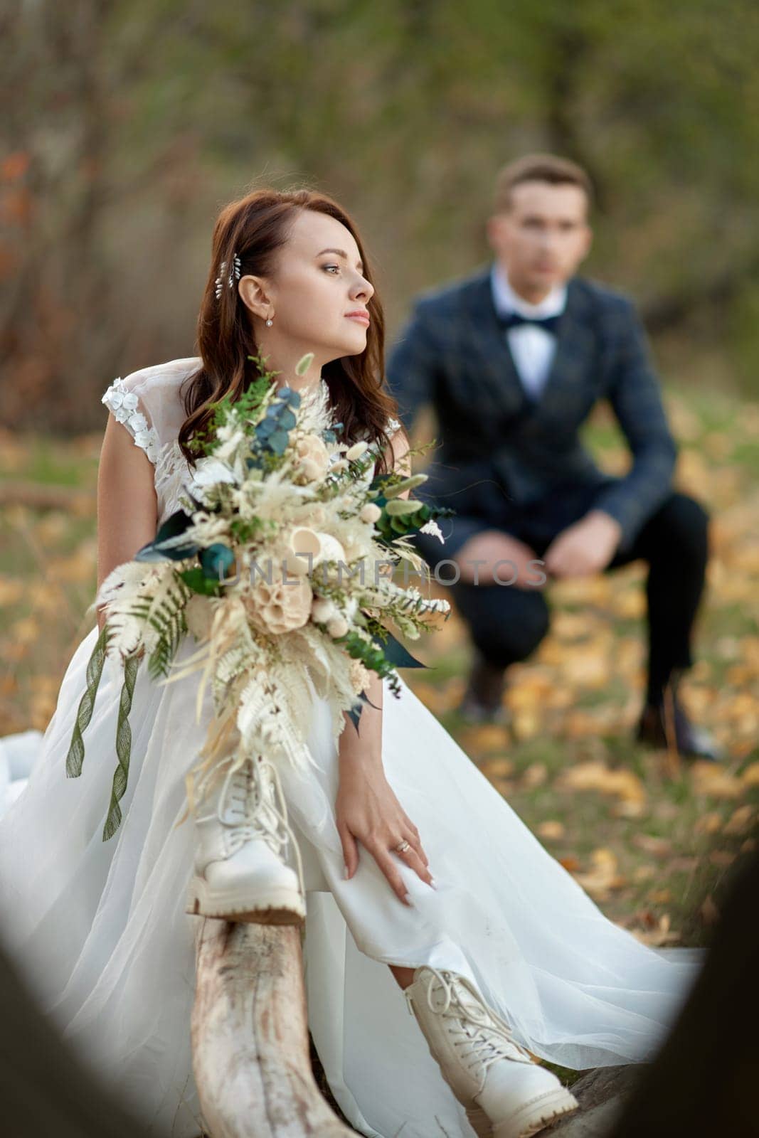 beautiful bride in white boots sitting on tree and holding autumn bouquet. blur groom on the background