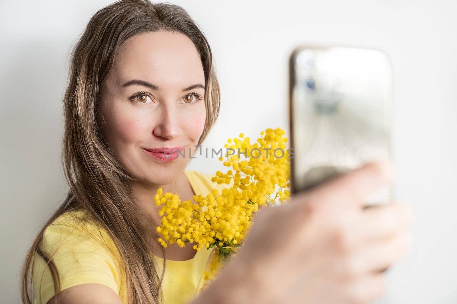 Beautiful young woman with mimosa flowers taking a selfie on white background.