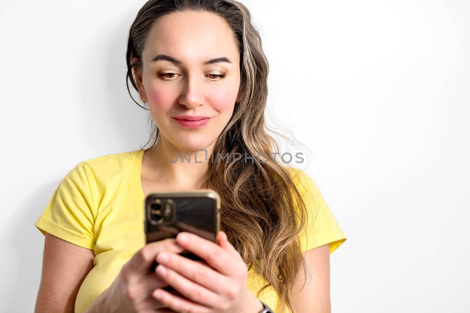 Smiling young woman in blank yellow t-shirt using smartphone on light background. Technology and people concept.
