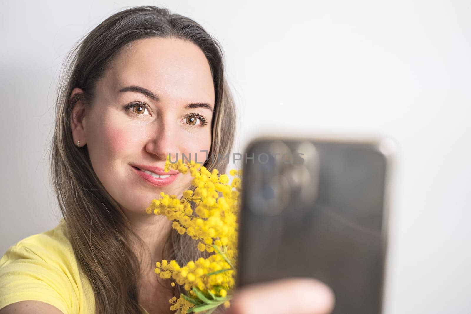 Beautiful young woman with mimosa flowers taking a selfie on white background.