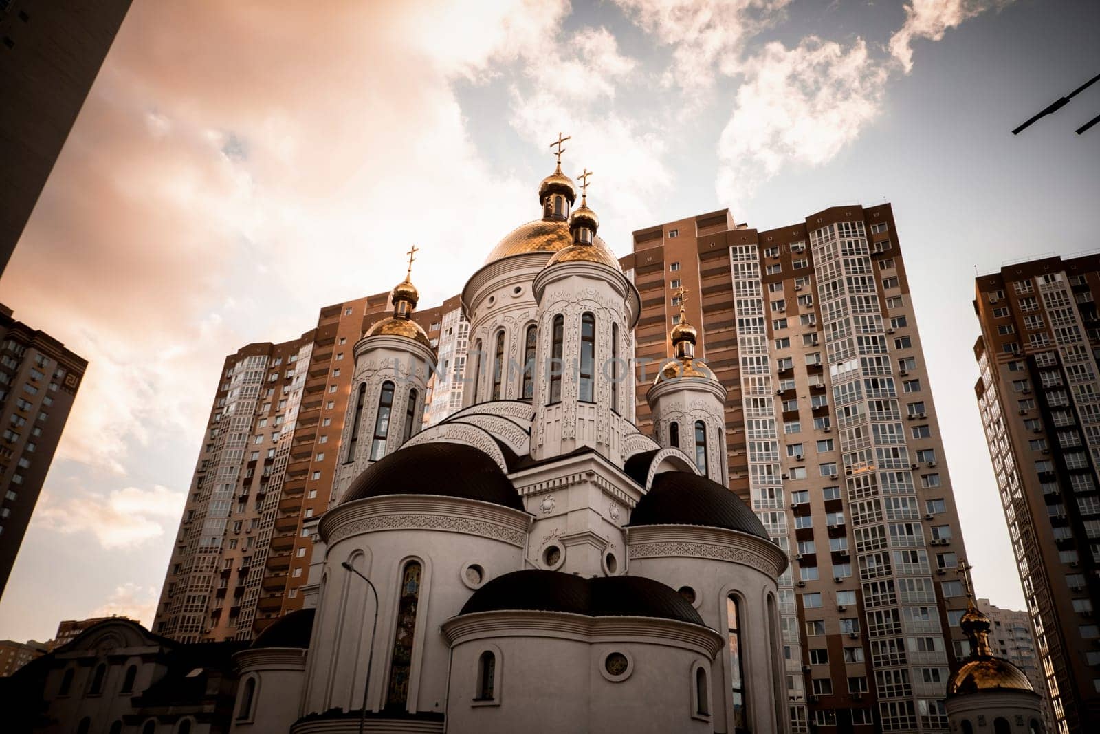 Church in town located in Kyiv. Bottom view of modern Church with skyscrapers on background, district against blue sky. Looking up at church buildings in downtown. Rising sun on the horizon. by igor010