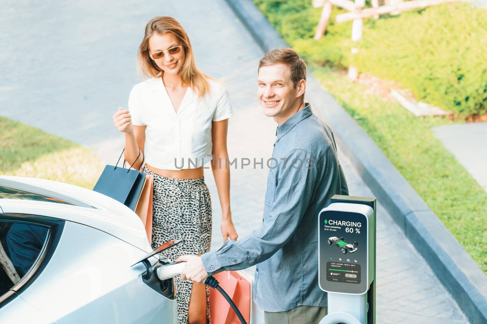 Young couple travel with EV electric car charging in green sustainable city outdoor garden in summer shows urban sustainability lifestyle by green clean rechargeable energy of electric vehicle innards