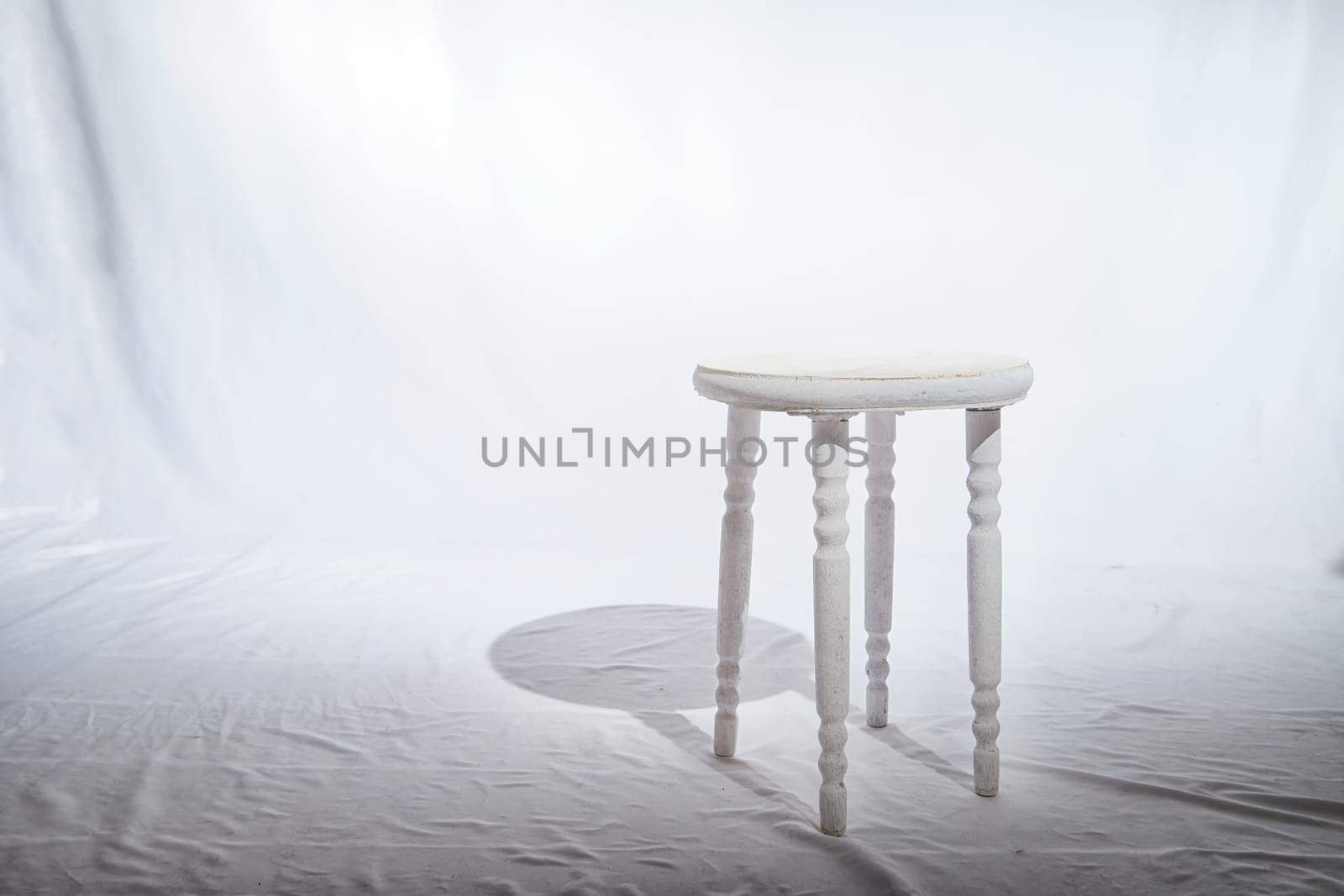 Solitary Wooden Stool in a White Minimalist Setting. Lone, white-painted wooden stool stands centered on textured surface, surrounded by soft white backdrop