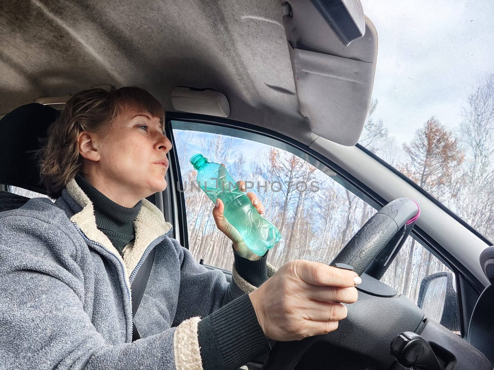 Portrait of female driver in solo journey. Adult mature woman holding steering wheel and bottle of water. Drinking while driving for to stay awake. Lady girl drinks alcohol vodka or gin while driving