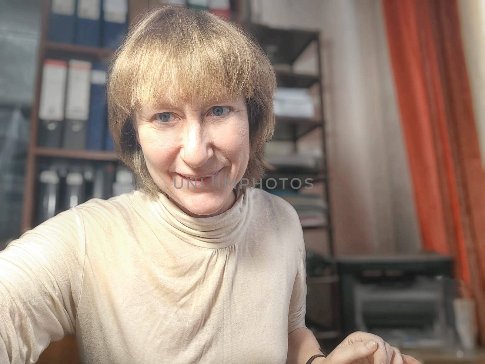 Funny blonde girl or a middle-aged woman poses for the camera with a smile and closet with folders on background. A relaxed Portrait of an Accountant, manager, blogger, freelancer in the workplace
