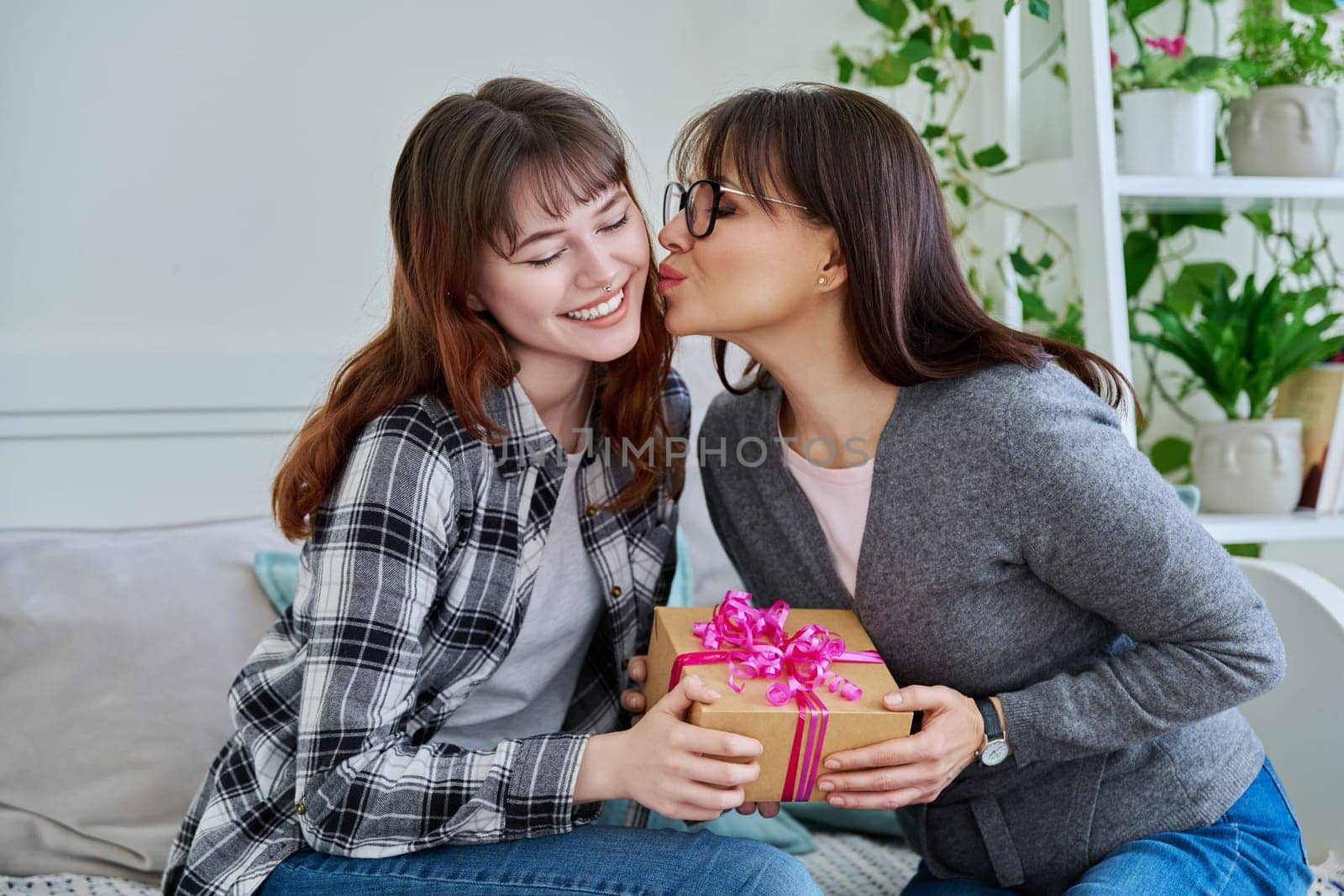 Cheerful teenage daughter congratulating middle aged mother with gift, sitting together on couch at home. Holiday birthday, congratulations, love, mother's day, happiness joy concept