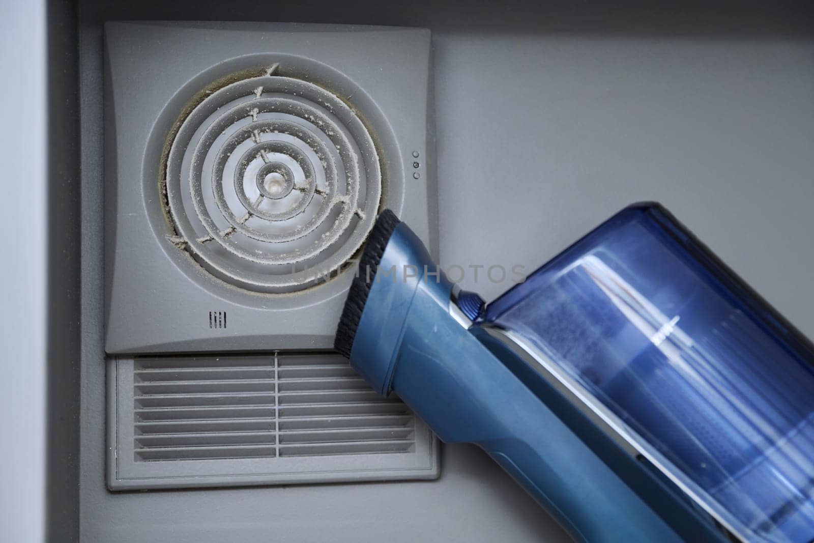 Cleaning wall-mounted dusty hood ventilation grill in bathroom with vacuum cleaner. Housekeeping, housework, housecleaning, cleaning service concept