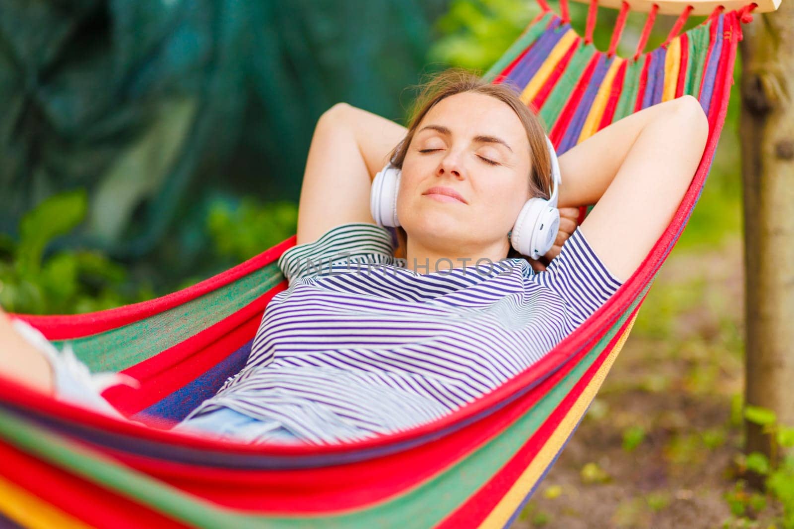 A serene woman lies in a colorful hammock outdoors, listening to music with white headphones, eyes closed in relaxation.