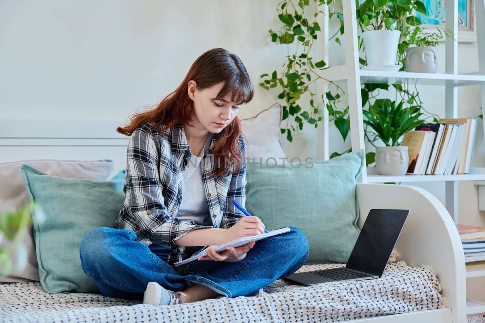 College student girl studying at home, sitting on the couch with laptop, making notes in study book. Education, training, technology, youth concept