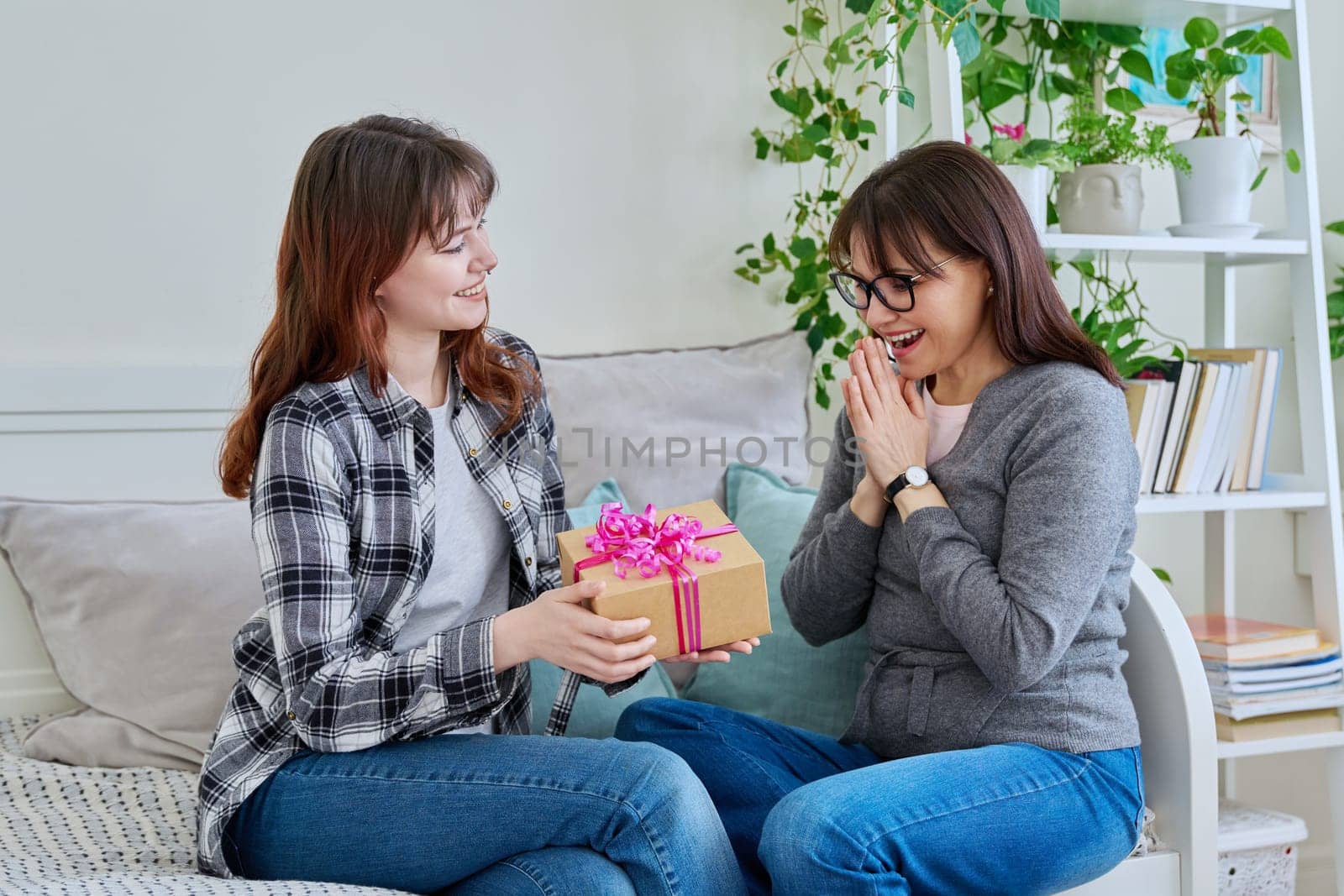 Cheerful teenage daughter congratulating middle aged mother with gift, sitting together on couch at home. Holiday birthday, congratulations, love, mother's day, happiness joy concept