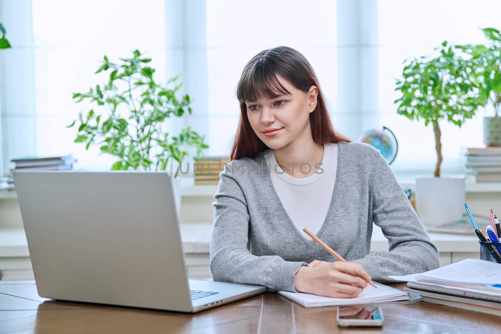 College student girl sitting at desk using laptop computer, making notes in study notebook, at home. Teenager female watching webinar, preparing for exam tests, studying remotely.