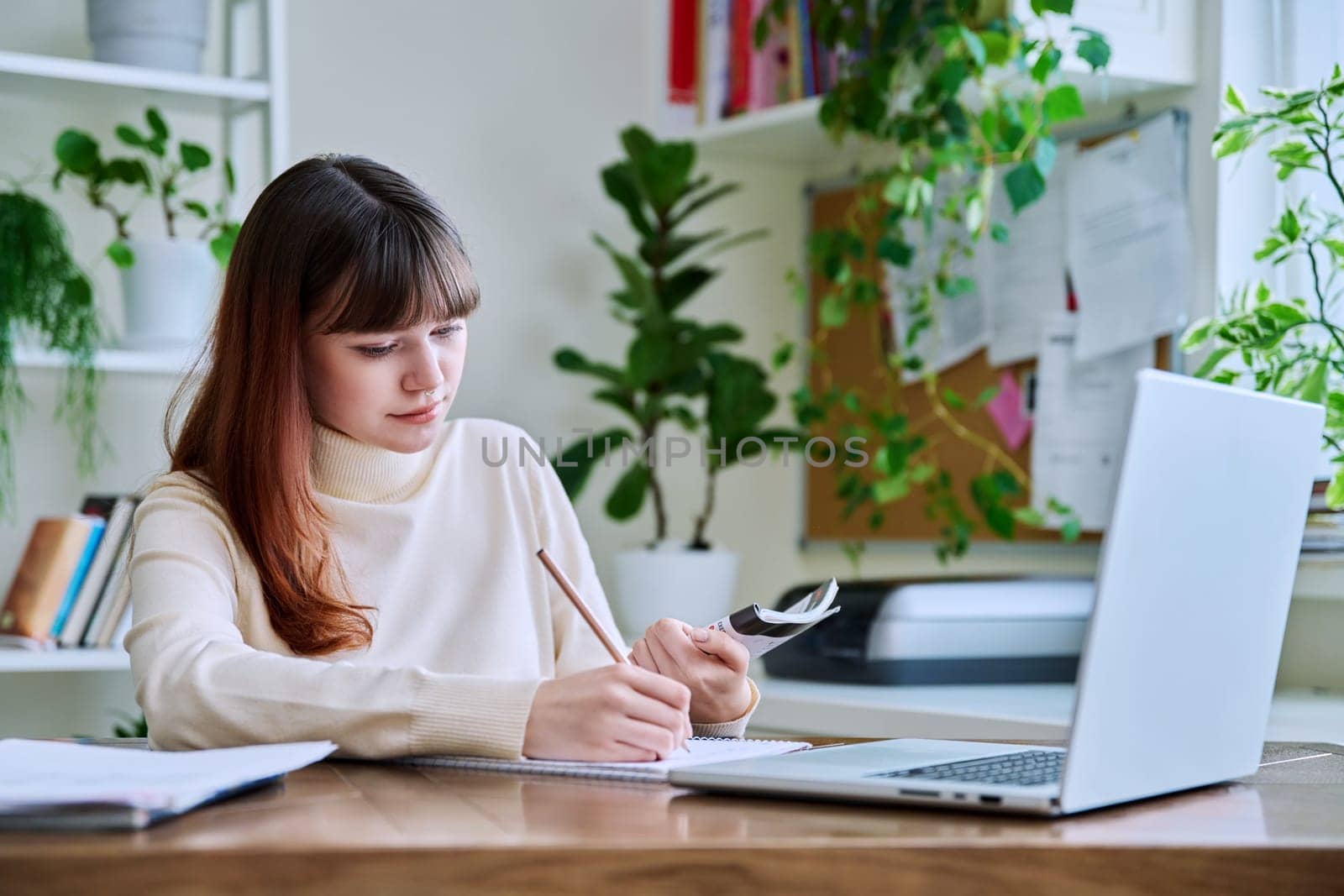 Young female college student studying at home at desk using computer laptop, writing in notebook. E-learning, education, technology, knowledge, youth concept