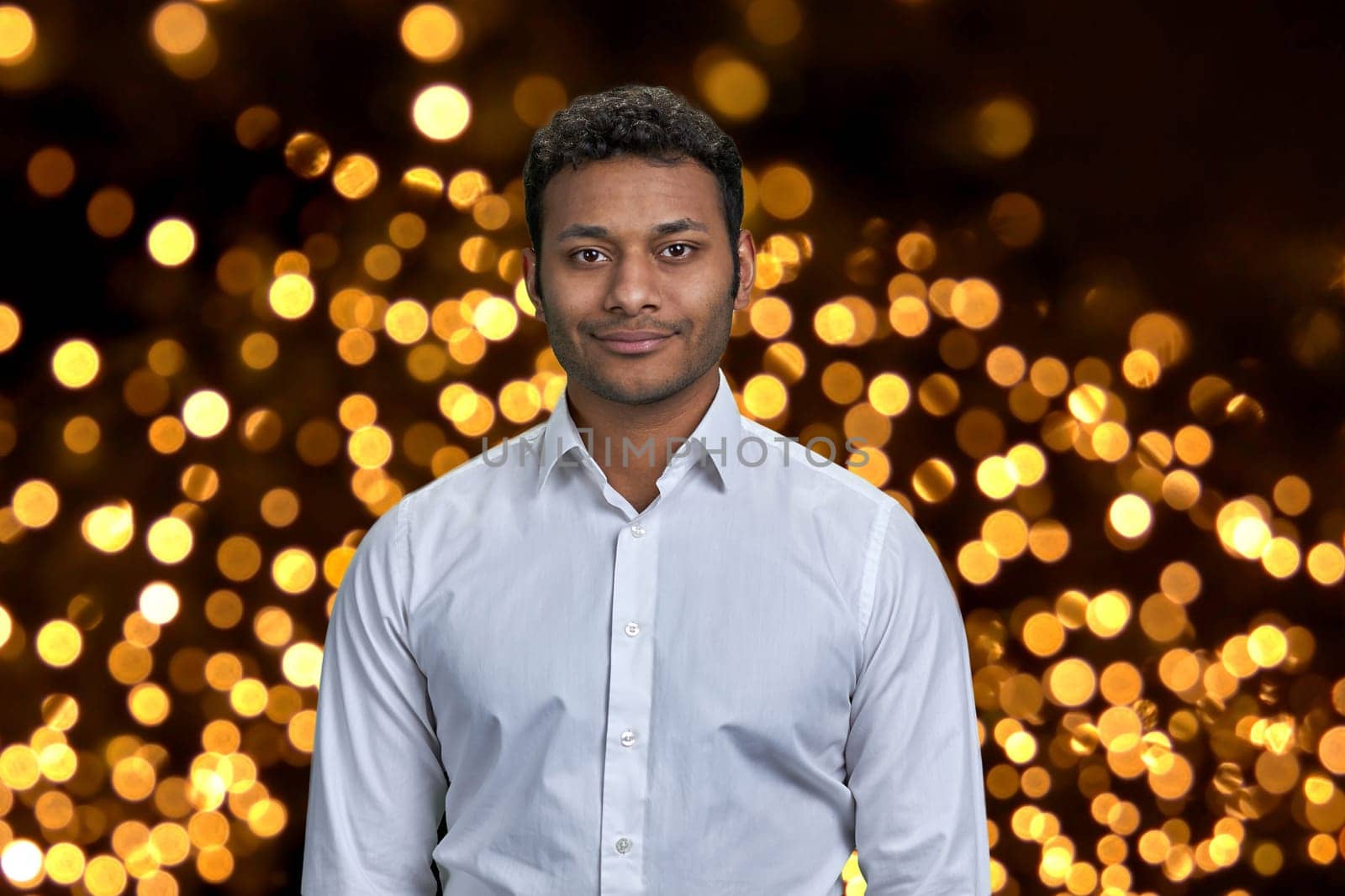 Handsome dark-skinned businessman wearing white shirt looking at camera. Black and golden bokeh lights in the background.