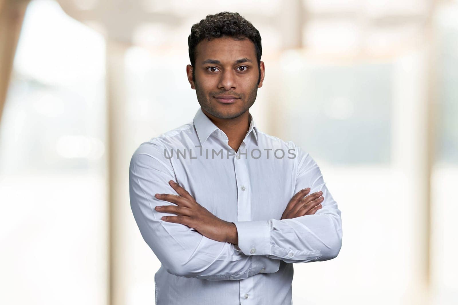 Attractive indian businessman looking at camera with crossed arms. Confident male executive standing on blur interior background.