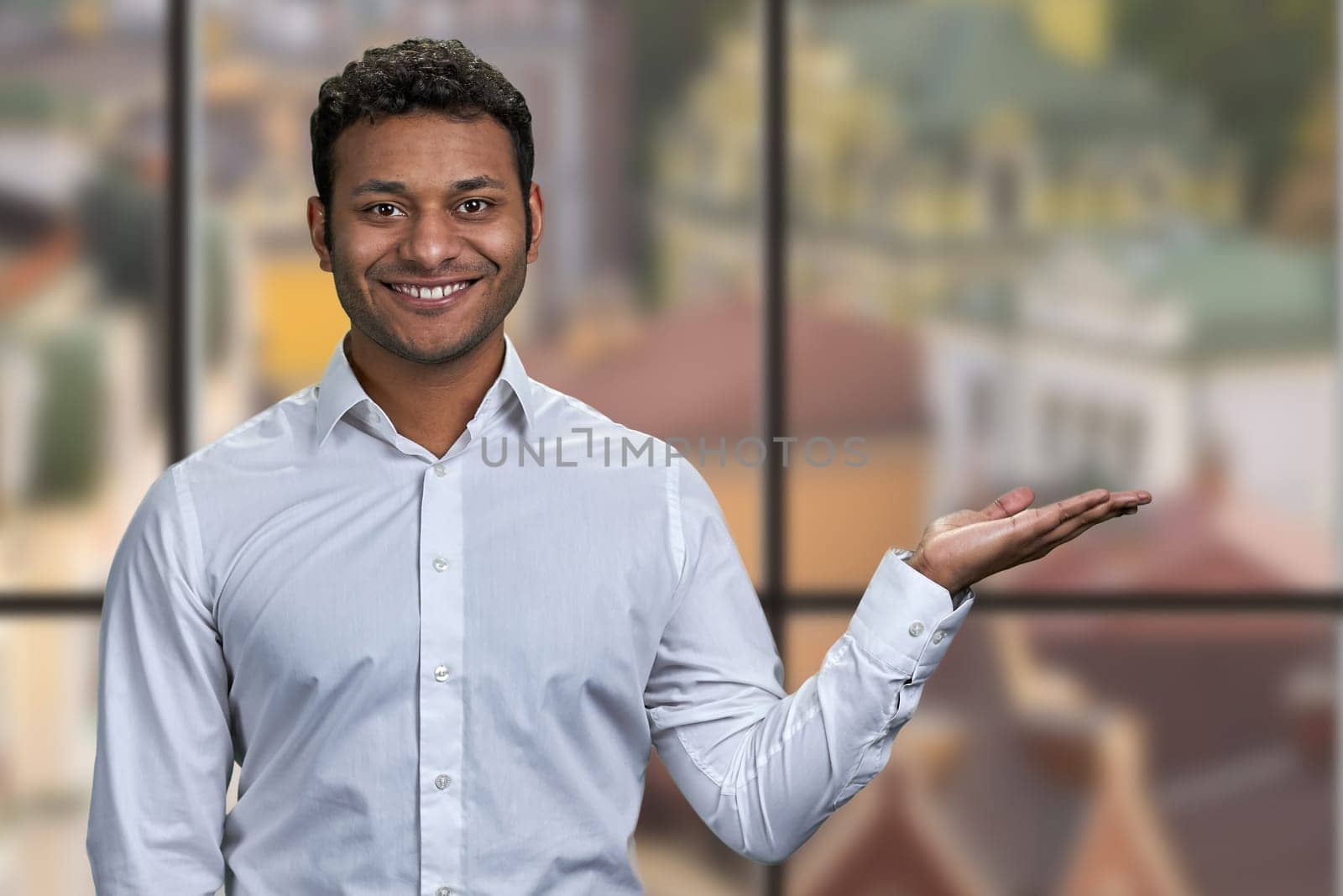 Handsome man in white shirt presenting something with open hand palm. Blur window background with view to city buildings.