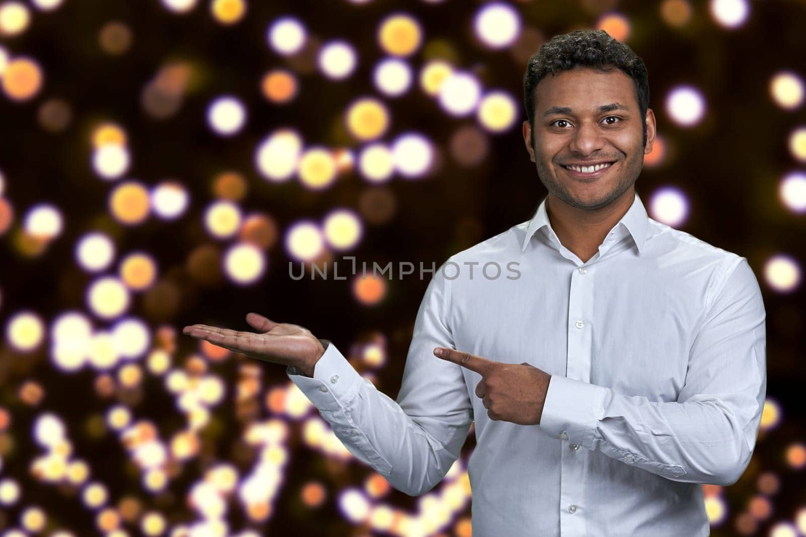 Handsome man looking at camera while presenting something on festive bokeh background. Christmas holiday deal.