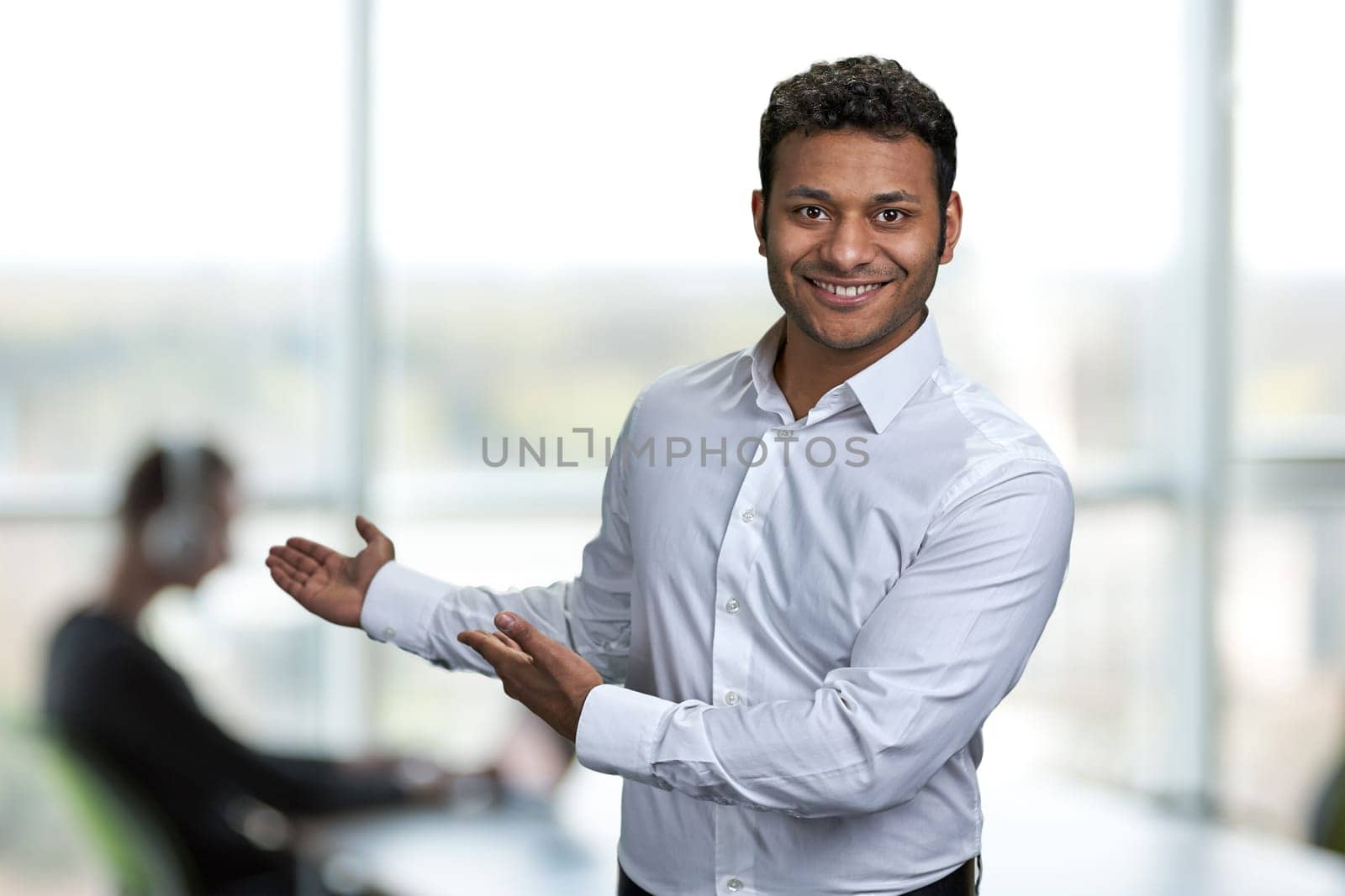 Young handsome man wearing white shirt pointing aside with hands open palms showing copy space. Attractive businessman presenting something on blurred office background.