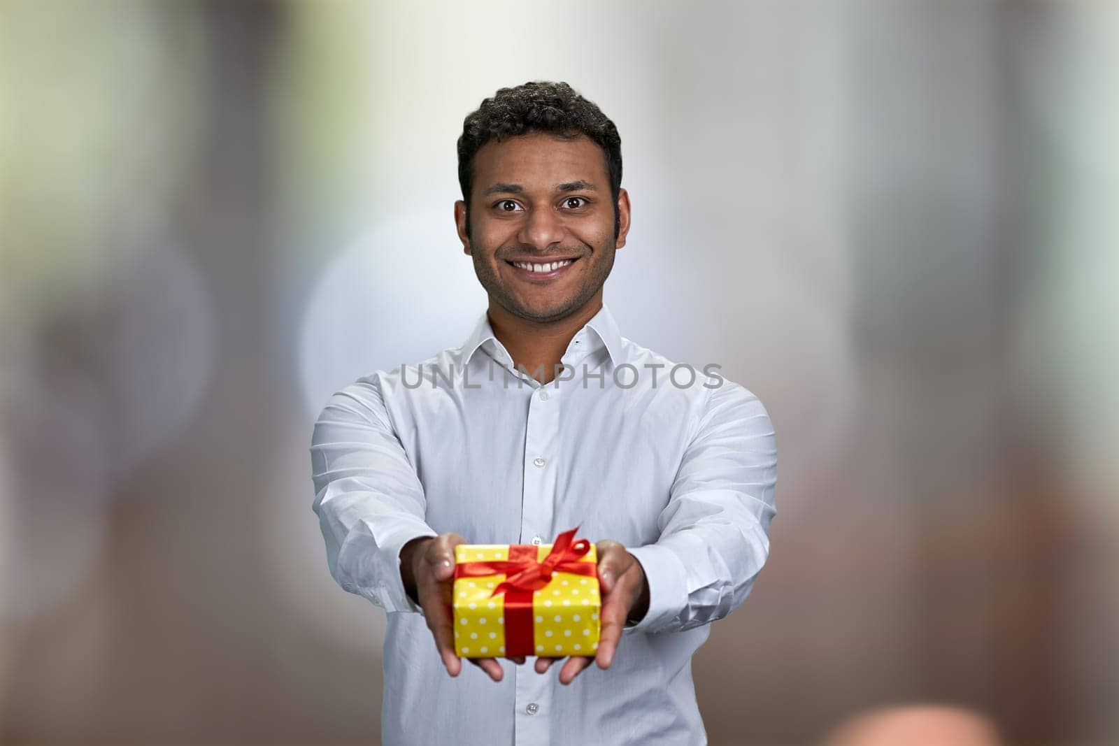 Young handsome businessman offering present box on abstract bokeh background. Birthday celebration concept.
