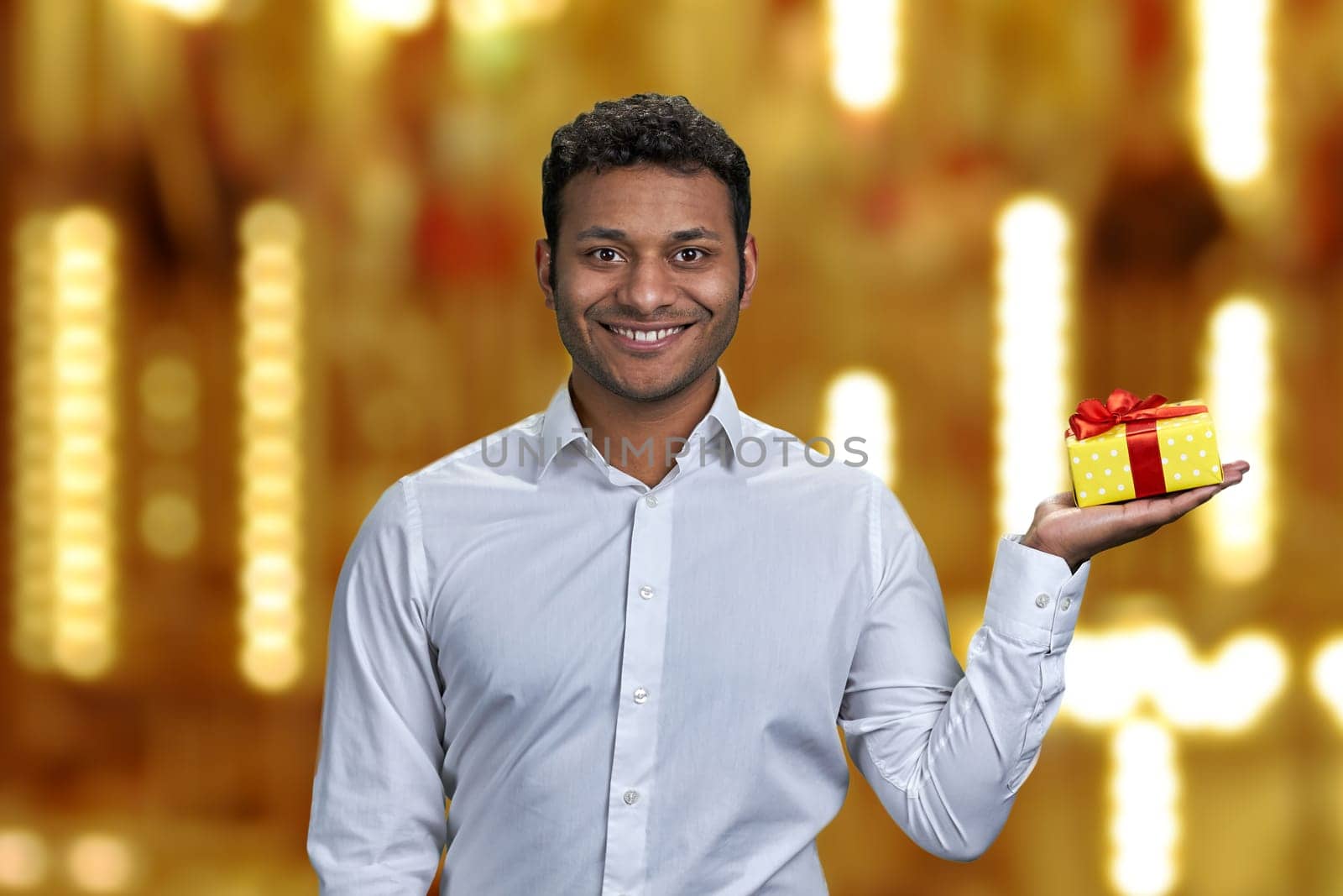 Young smiling man holding gift box on his palm and looking at camera. Time for Christmas gifts.