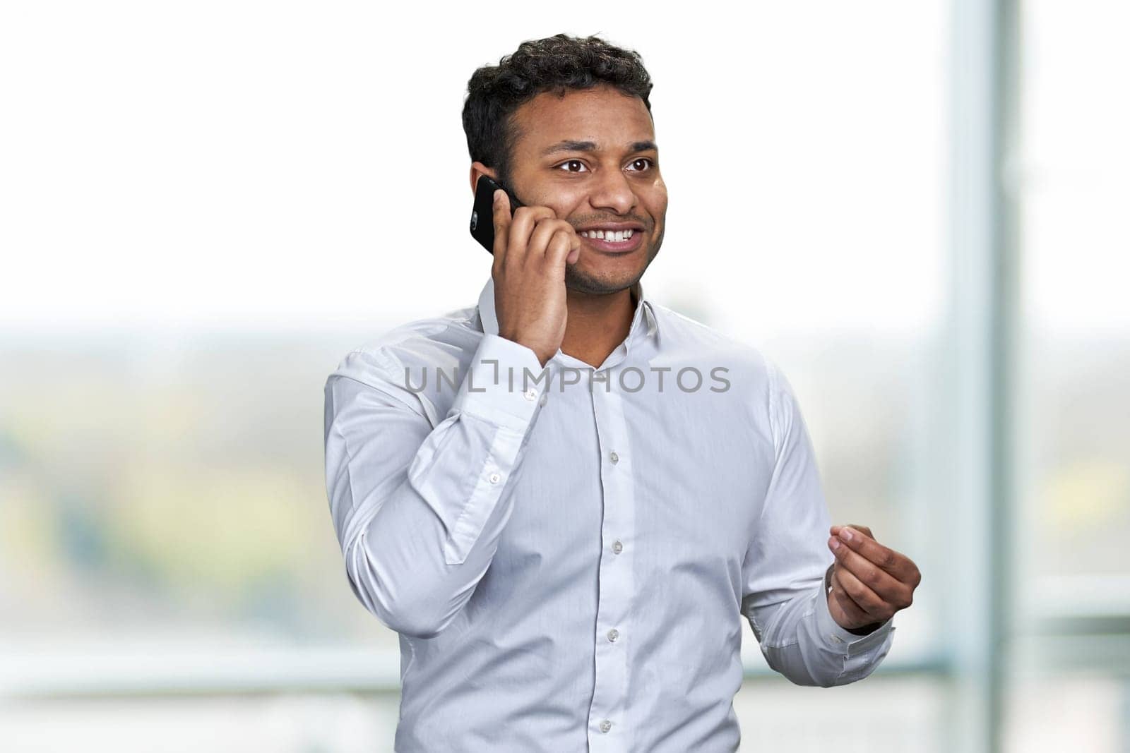 Cheerful young businessman wearing white shirt talking on mobile phone. Blur interior background.