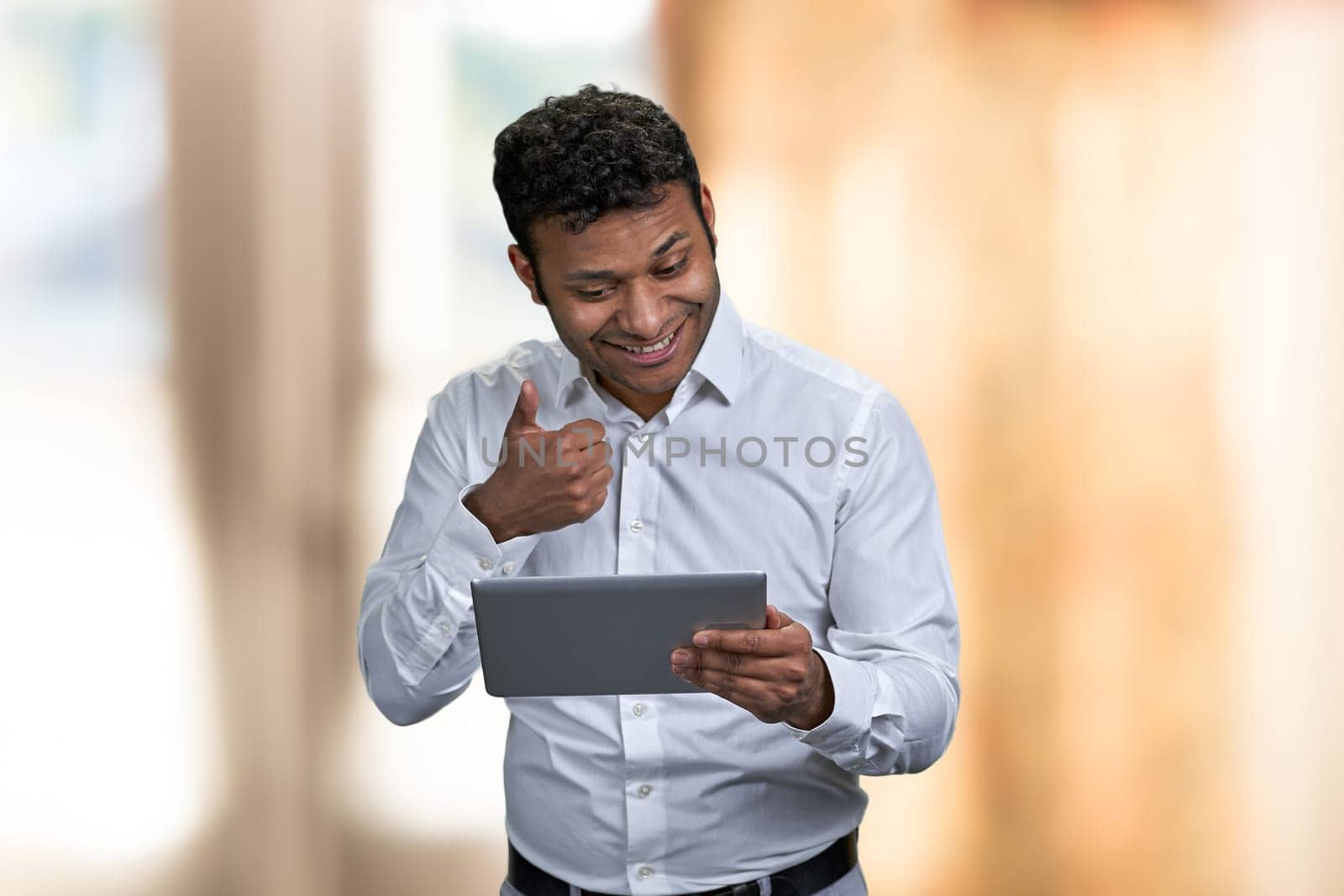 Handsome young businessman showing thumb up while having video call on digital tablet. Blur interior background.