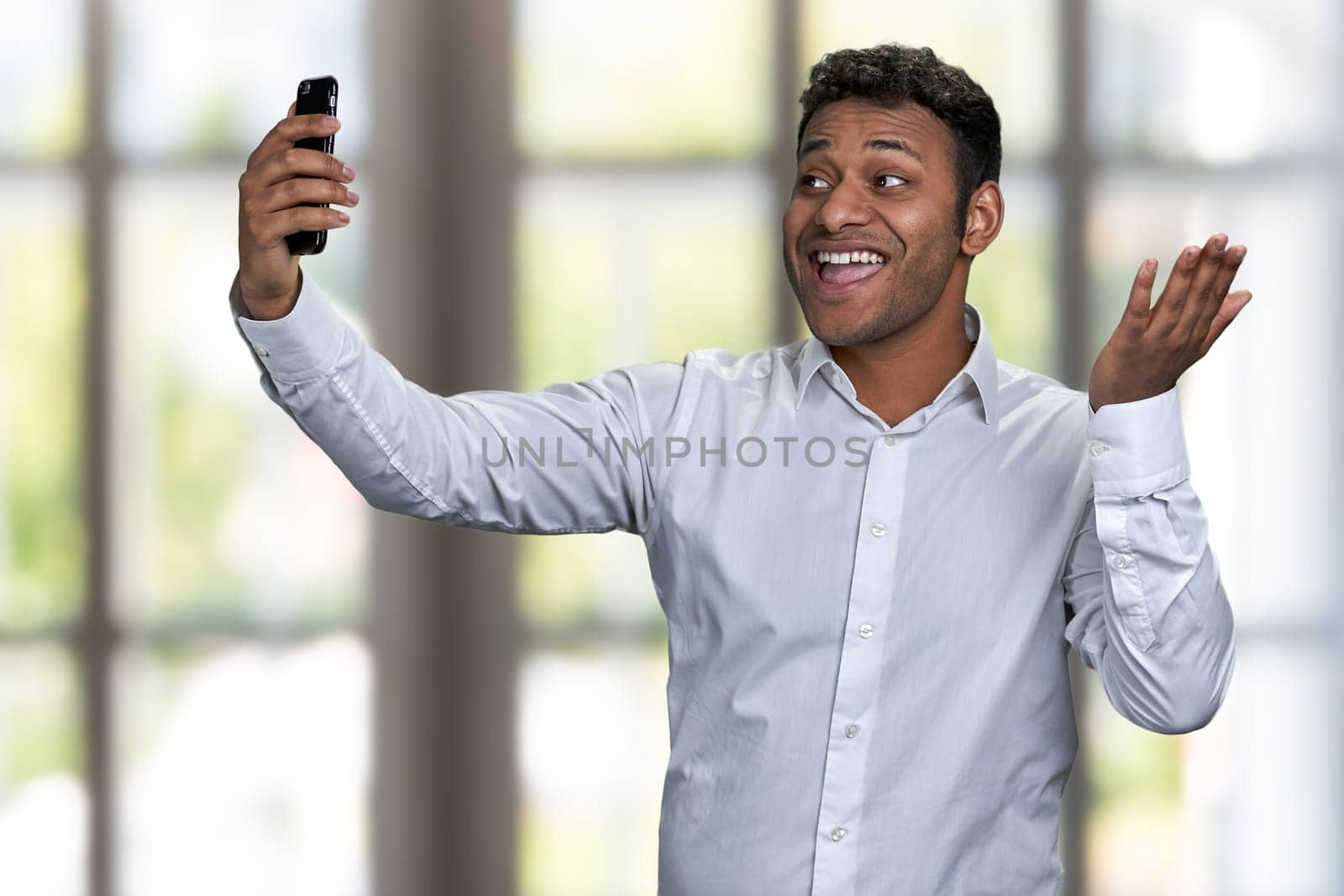 Attractive cheerful indian man in white shirt using cell phone on blur interior background. People, technology and fun concept.