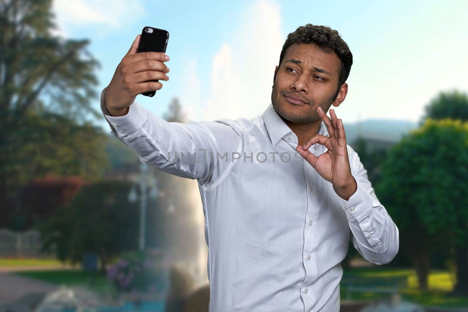 Handsome young man showing okey gesture while taking selfie outdoors. People, modern technology, gestures and lifestyle concept.
