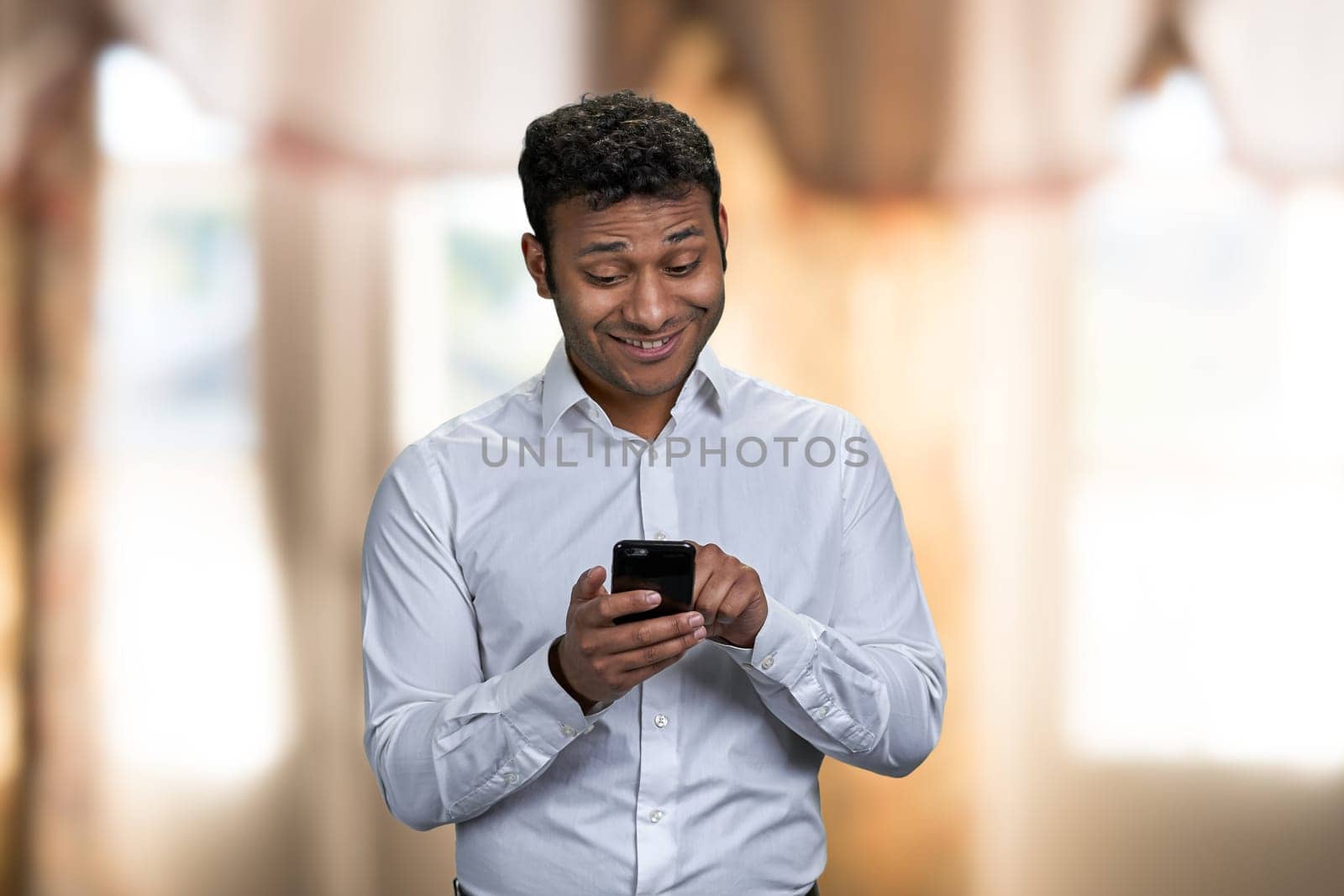 Handsome young man in white shirt surfing internet on mobile phone. Cheerful indian man using cell phone on blur interior background.