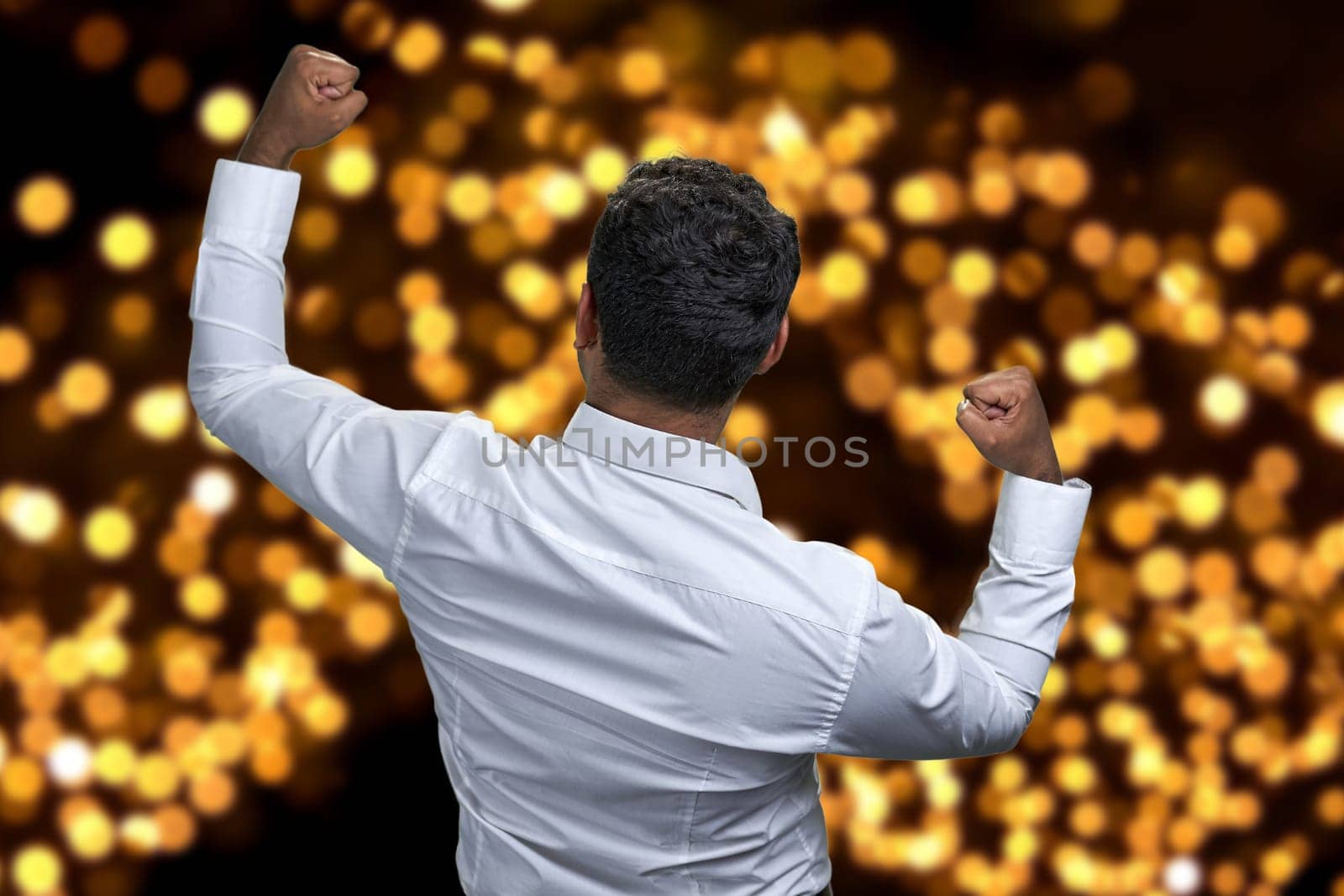 Rear view of successful businessman with clenched fists celebrating goal achievement. Festive bokeh lights in the background.