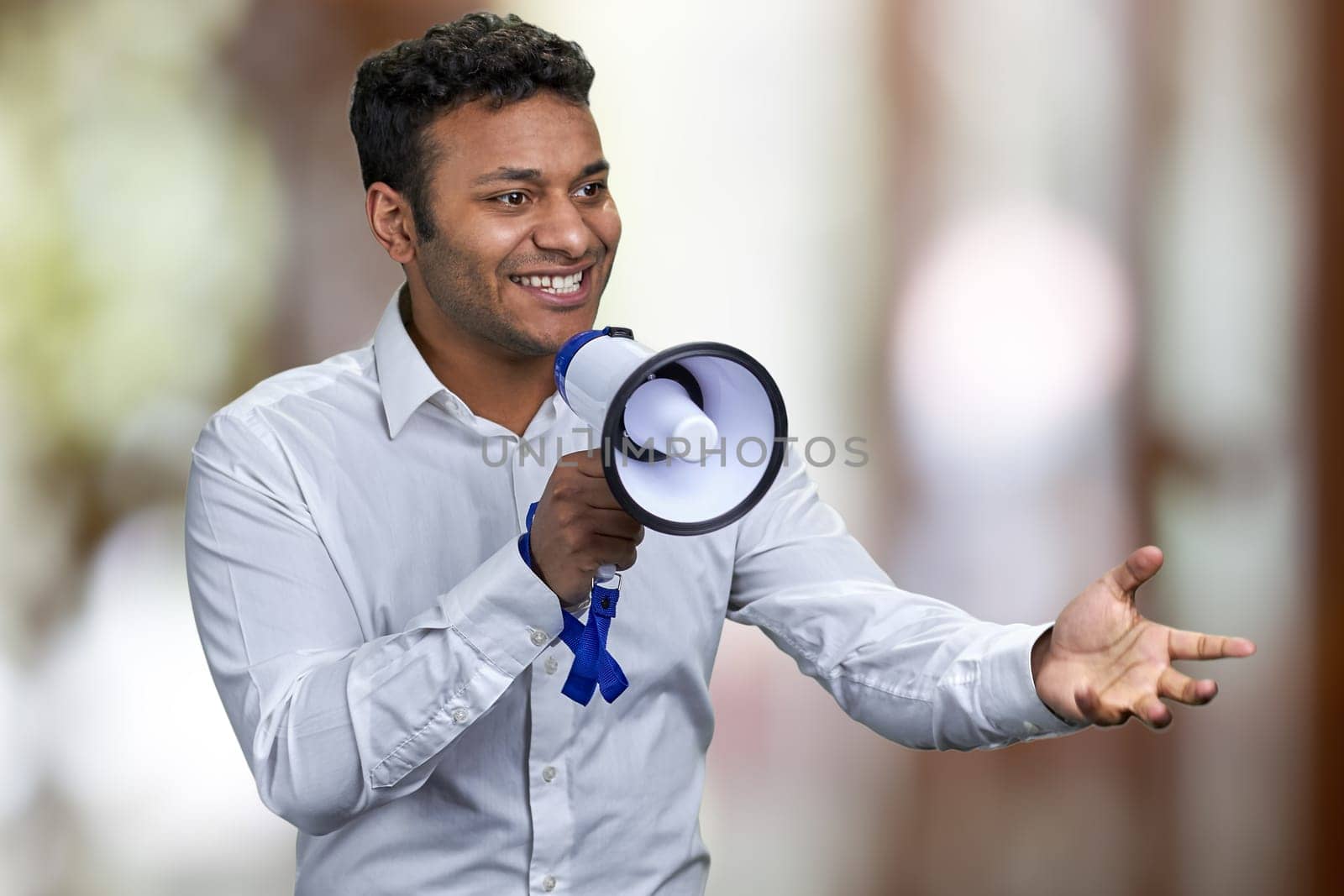 Cheerful young man in white shirt talking into megaphone. Abstract bokeh background.