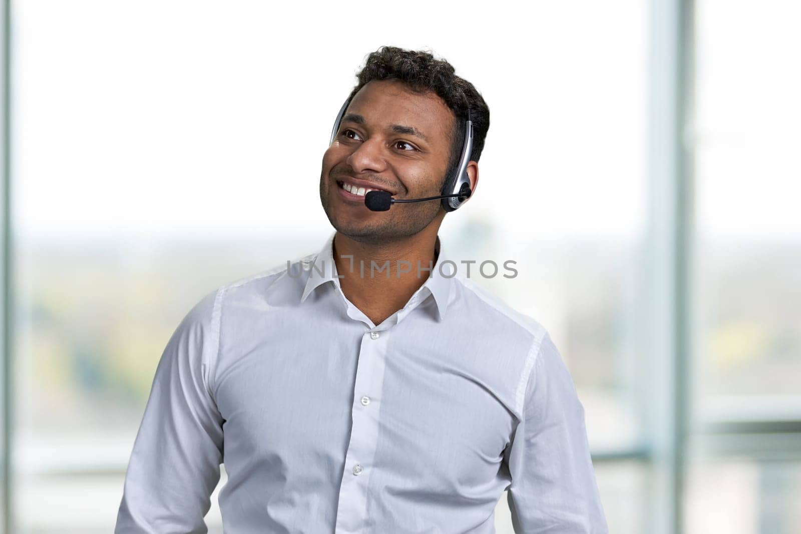 Cheerful male call center operator on blur interior background. Friendly smiling call center agent looking aside.