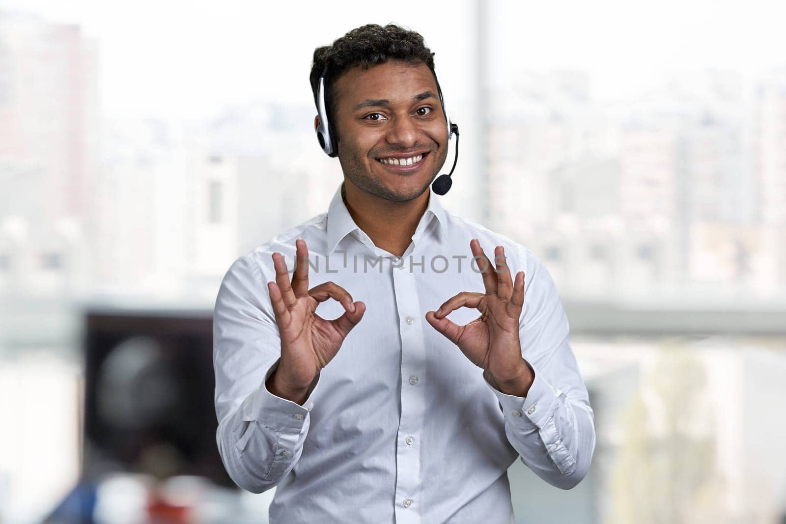 Cheerful male call center operator gesturing okey with both hands. Blur office interior in the background.