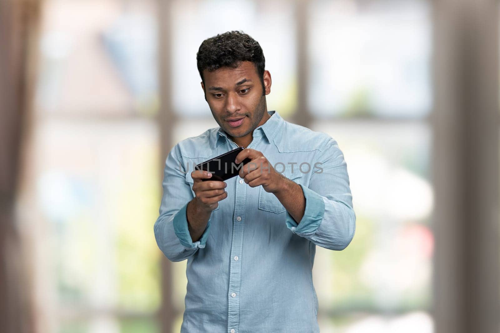 Attractive young man playing game on his smartphone. Blur interior background.
