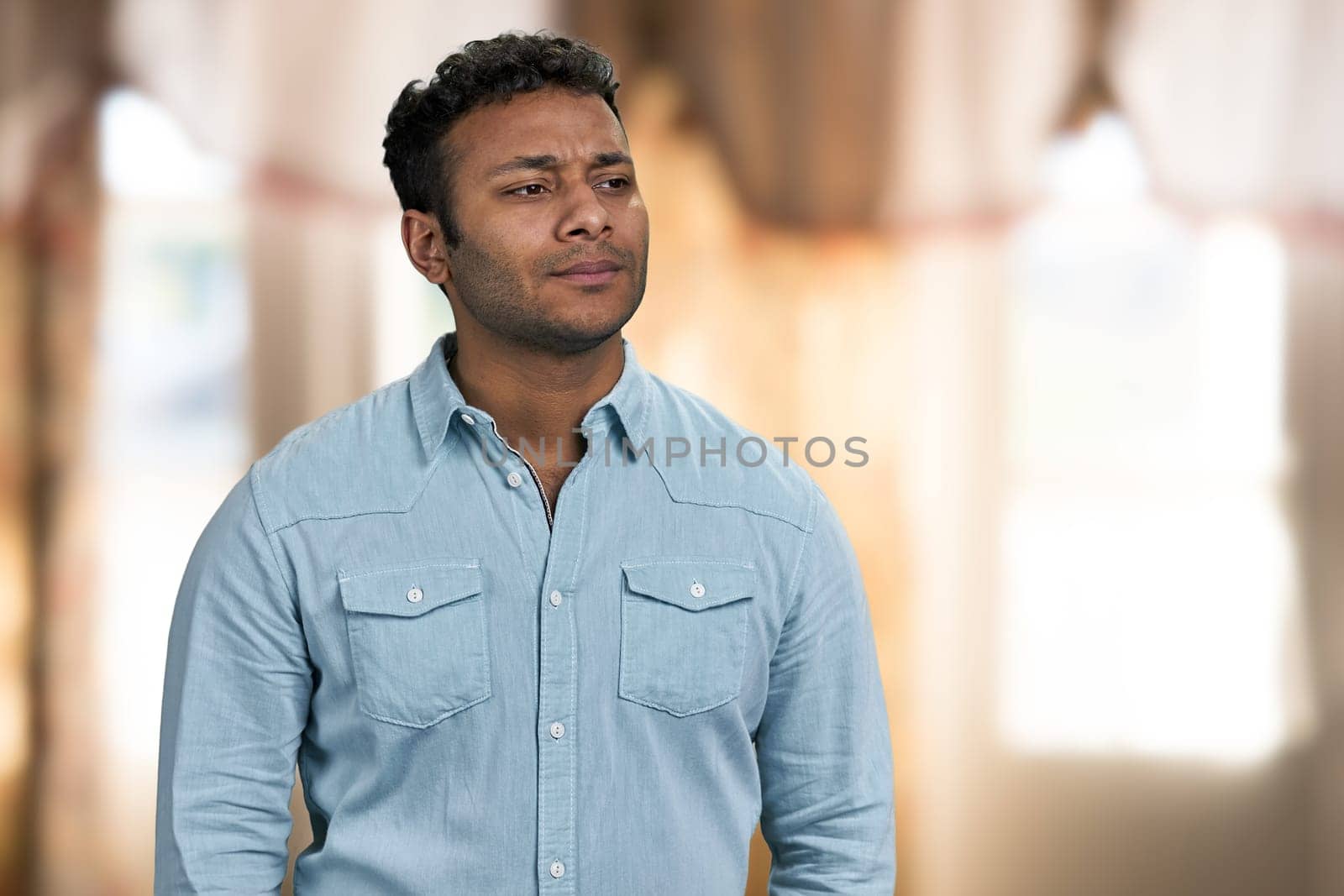 Young thoughtful man standing on interior blur background. Serious pensive man looking aside.