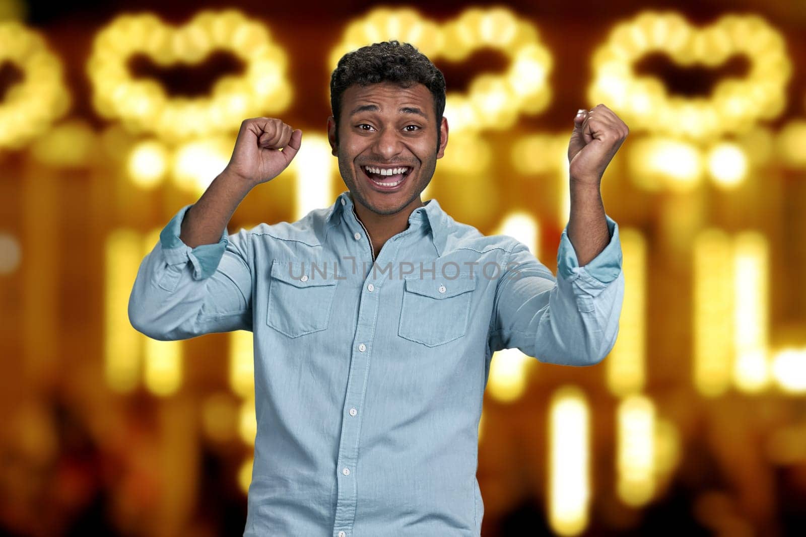 Young excited man celebrating success with clenched fists. Golden bokeh lights in the backgorund.