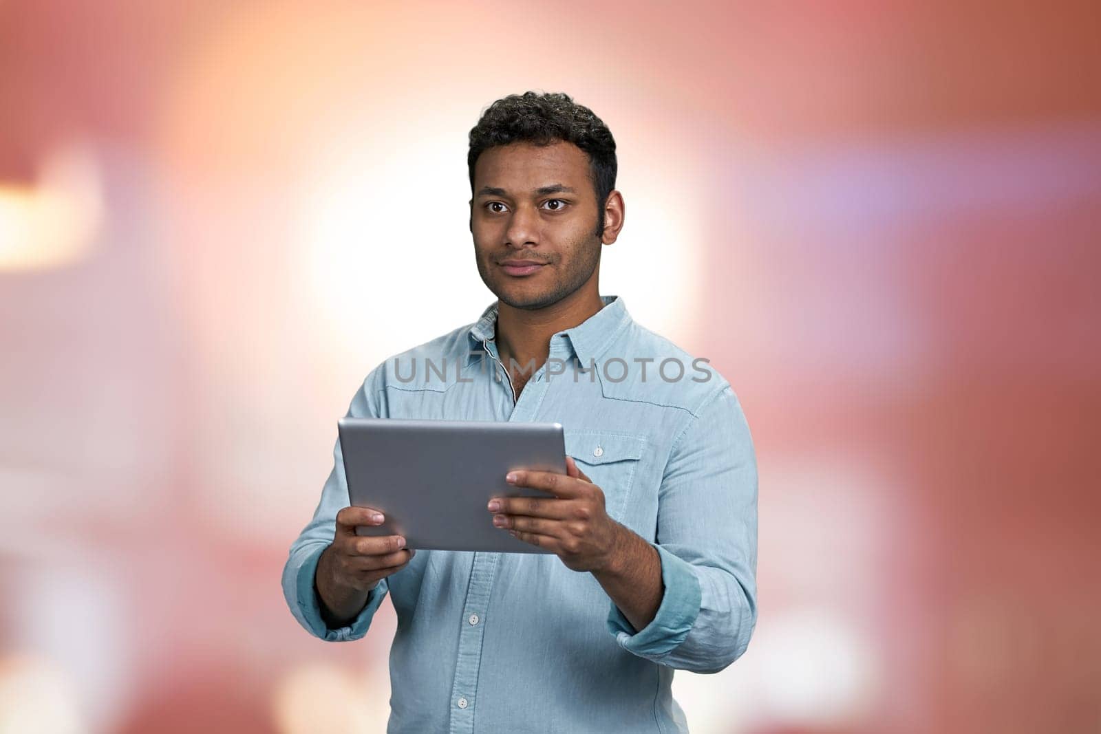 Young indian man using digital tablet on abstract pink bokeh background. People, technology and lifestyle concept.