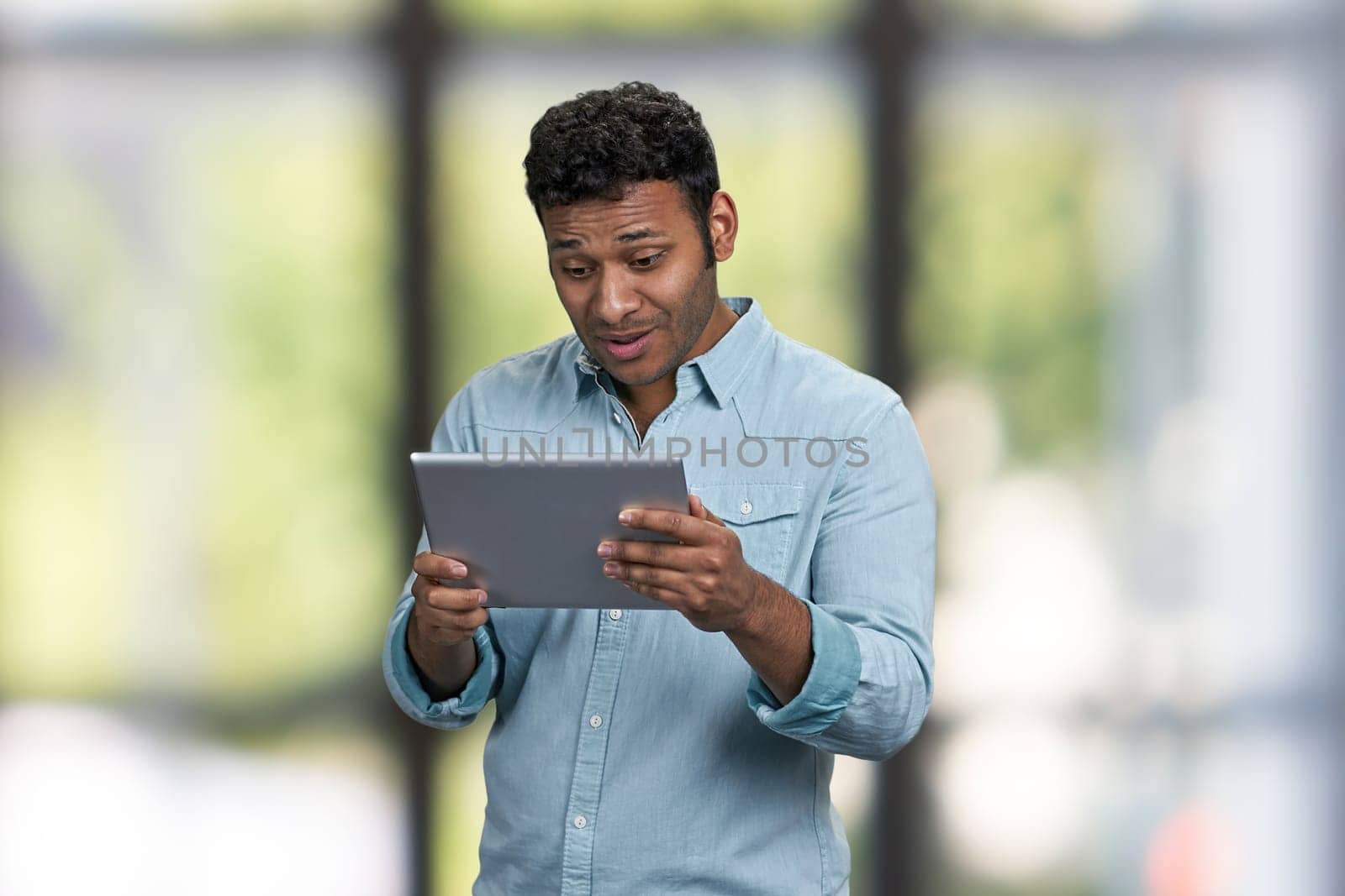 Surprised young man holding digital tablet on interior blur background. People, modern technology and lifestyle concept.