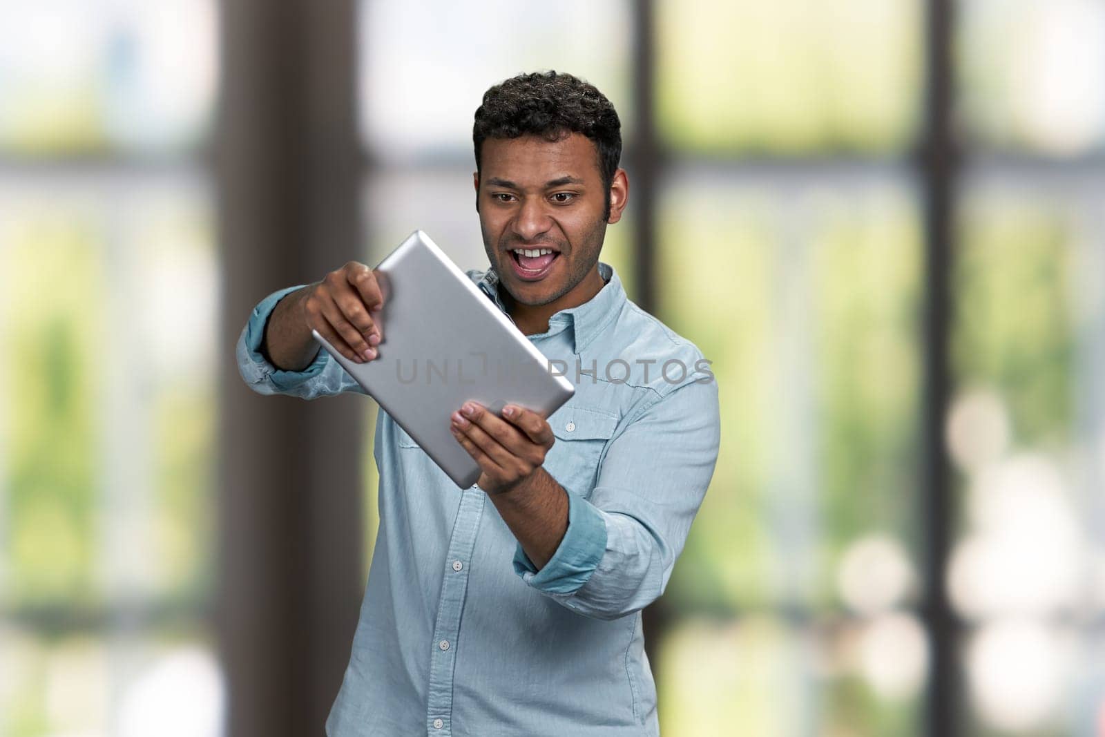 Energetic young man wearing blue shirt playing game on computer tablet. Interior blur background.