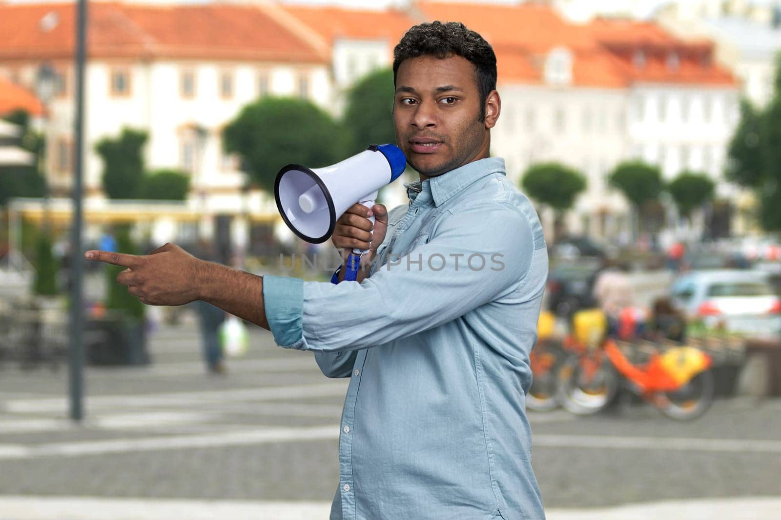 Young indian man talking into megaphone and pointing with index finger. Blur city street in the background.