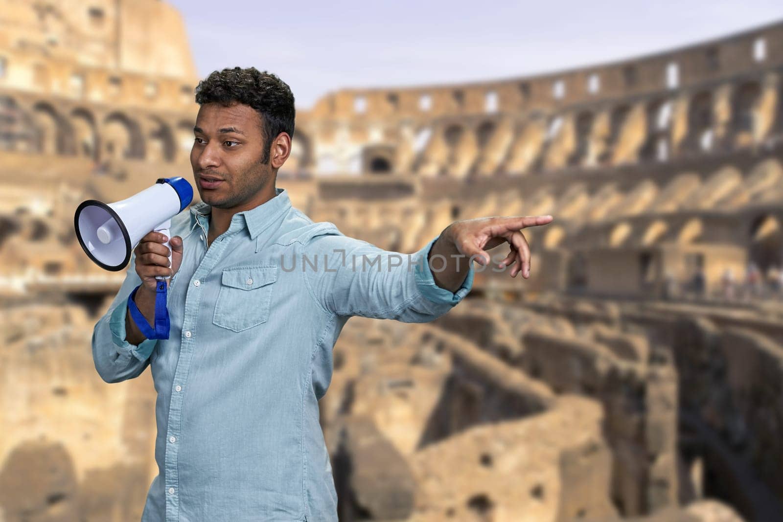 Young indian man talking into megaphone while standing on blurred background of Colosseum at Rome, Italy. People, tourism, ancient architecture and history concept.