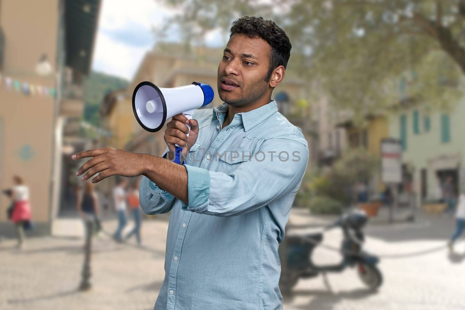 Young man with megaphone standing on blur city street background. Indian man with loudspeaker outdoors.